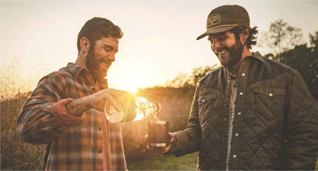Two men outdoors at sunset, raising glasses of Dos Primos tequila together.