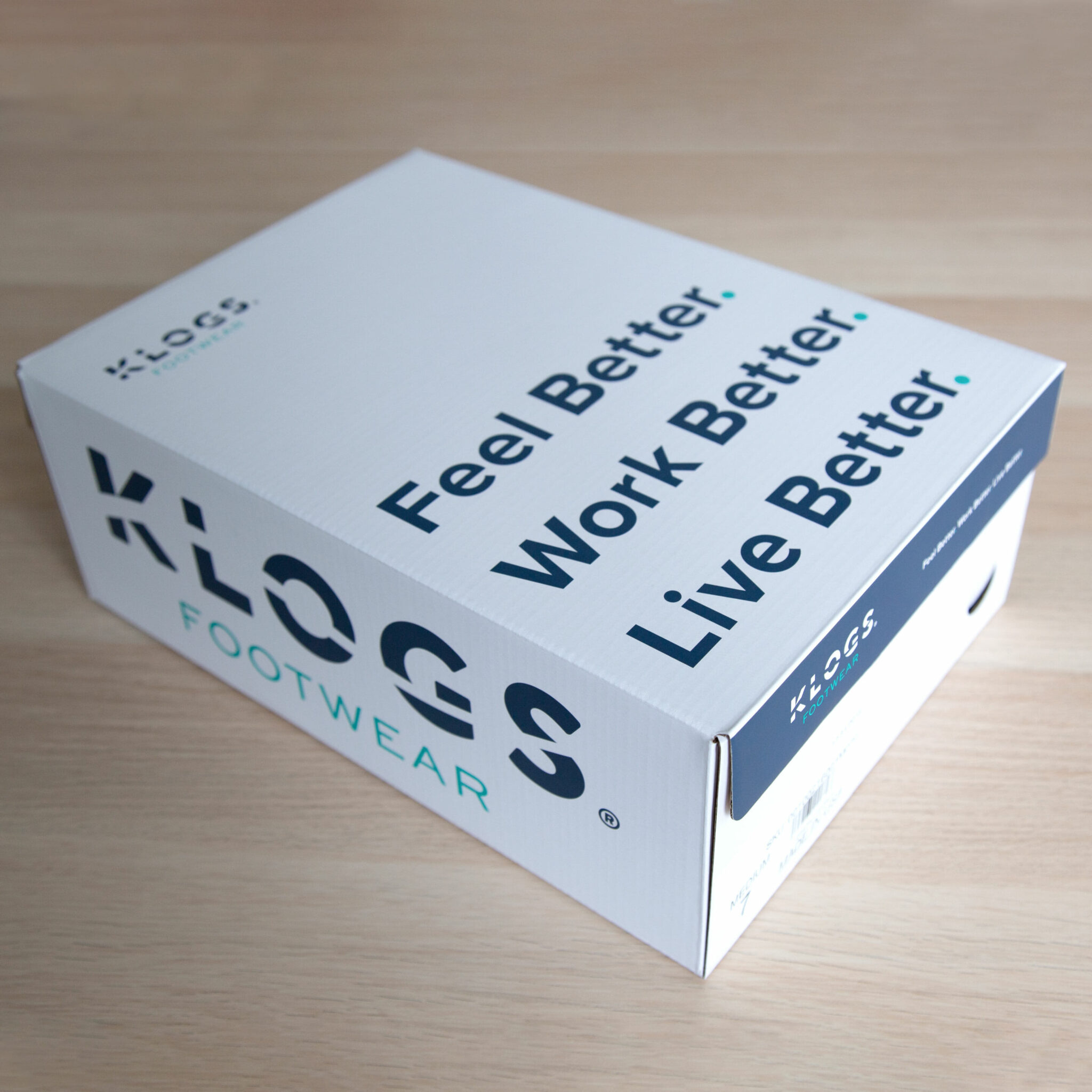 The Klogs Footwear shoebox on a pale wood floor, the lid printed with the KLOGS Footwear wordmark and the line “Feel Better. Work Better. Live Better.”
