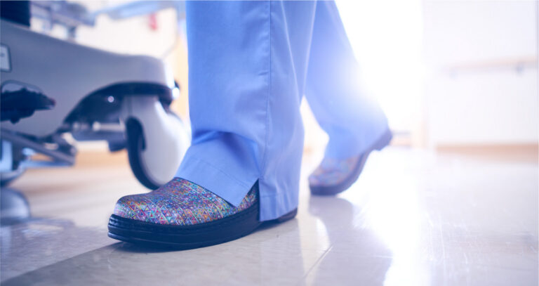 A nurse in blue scrubs walking a hospital corridor in glittered Klogs clogs.