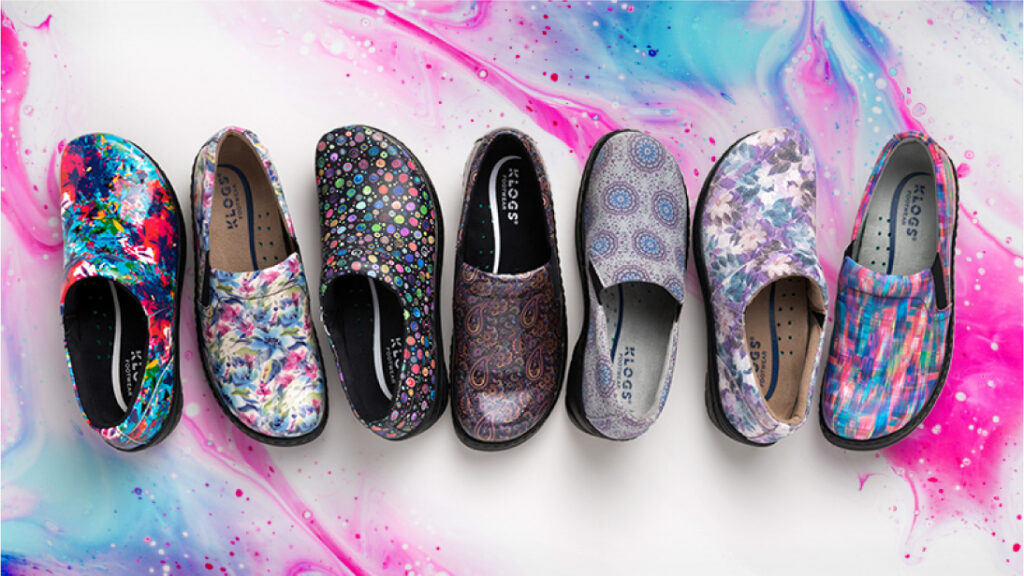 Seven pairs of Klogs clogs in a row, from painted floral patterns to plain grey, on a pink and blue marbled surface.